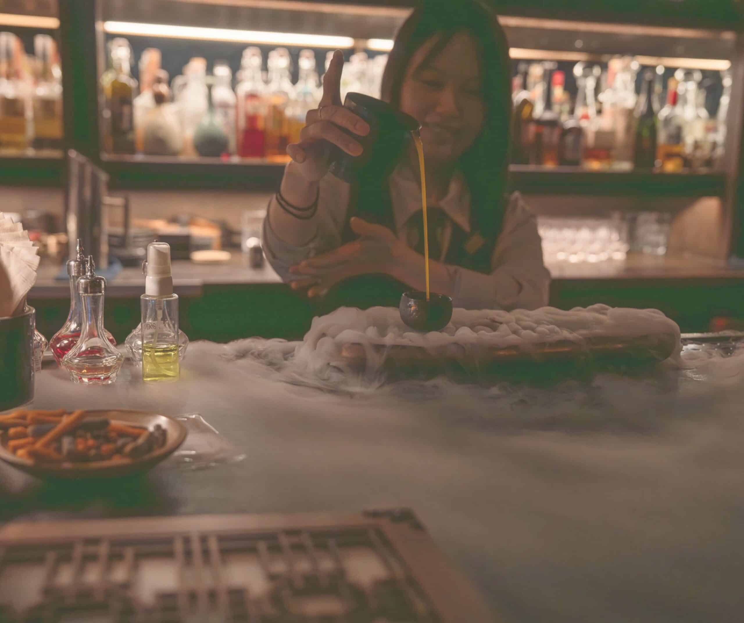 Bartender pouring a tall stream of yellow liquid into a glass at a dimly lit bar, bottles lined up in the background (blurred).