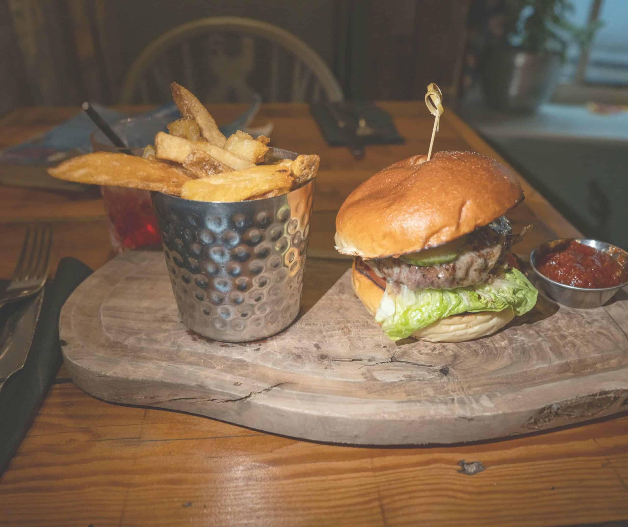 Burger with lettuce and pickles served with a stainless steel cup of chunky fries and chili jam on a wooden board at Hoof in Rye