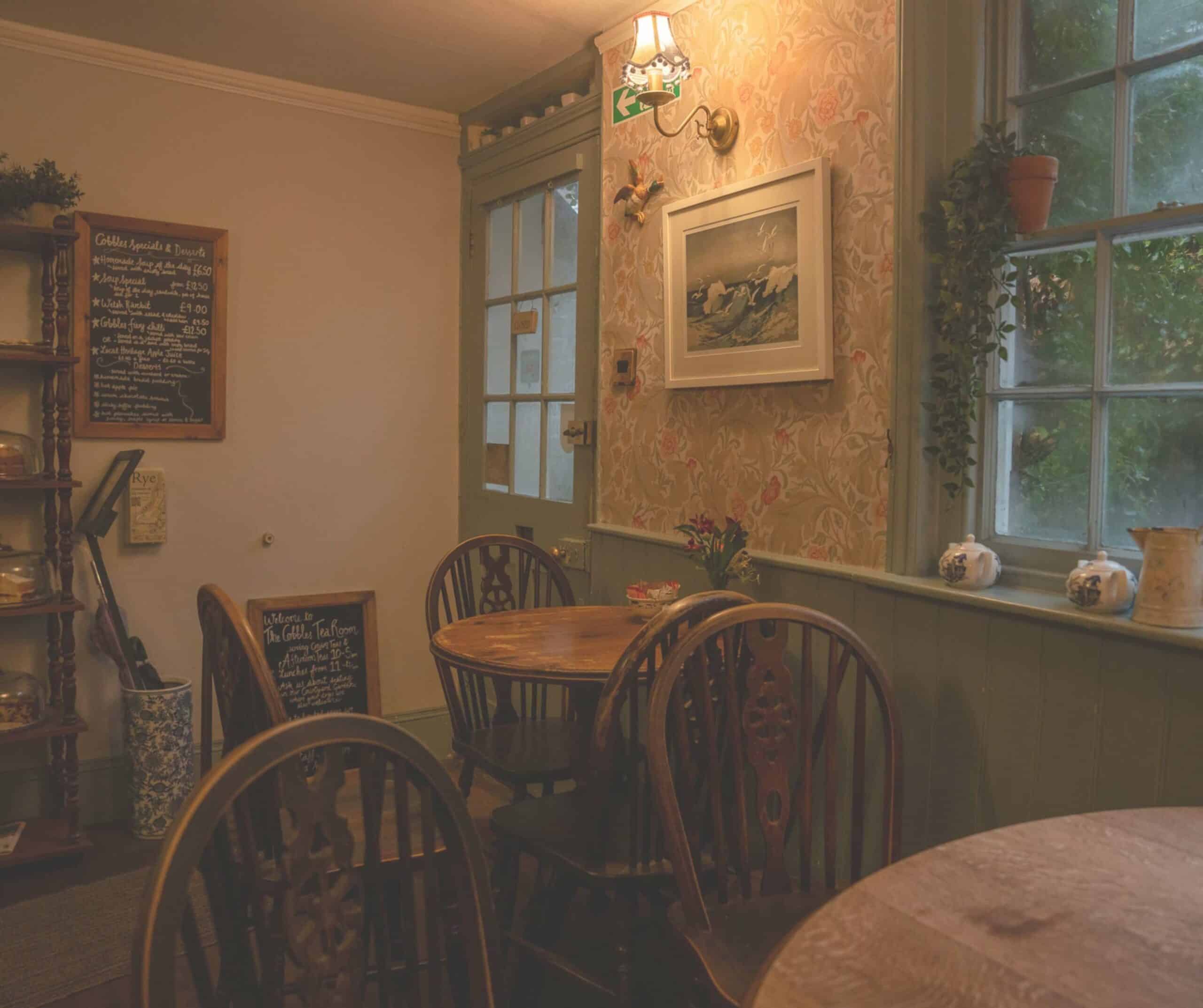 Seating area at The Cobbles Tea Room in Rye with floral wallpaper, Windsor chairs, teapots on the windowsill, and a garden view
