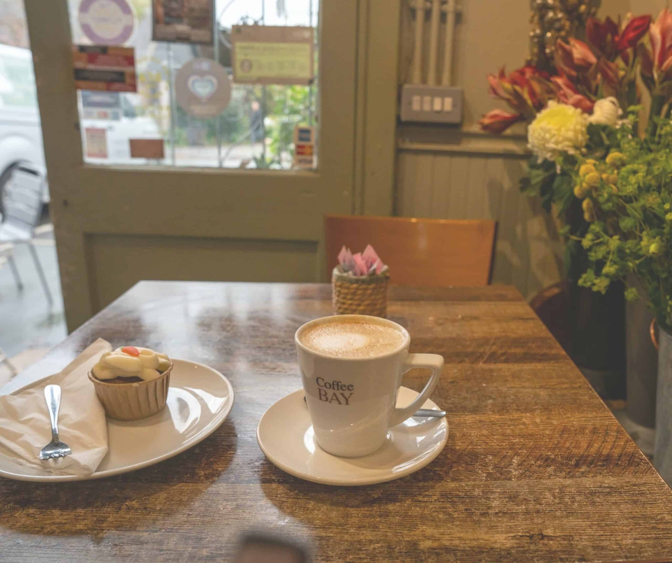 Cappuccino and a carrot cake cupcake on a wooden table surrounded by fresh flowers at Café des Fleurs in Rye