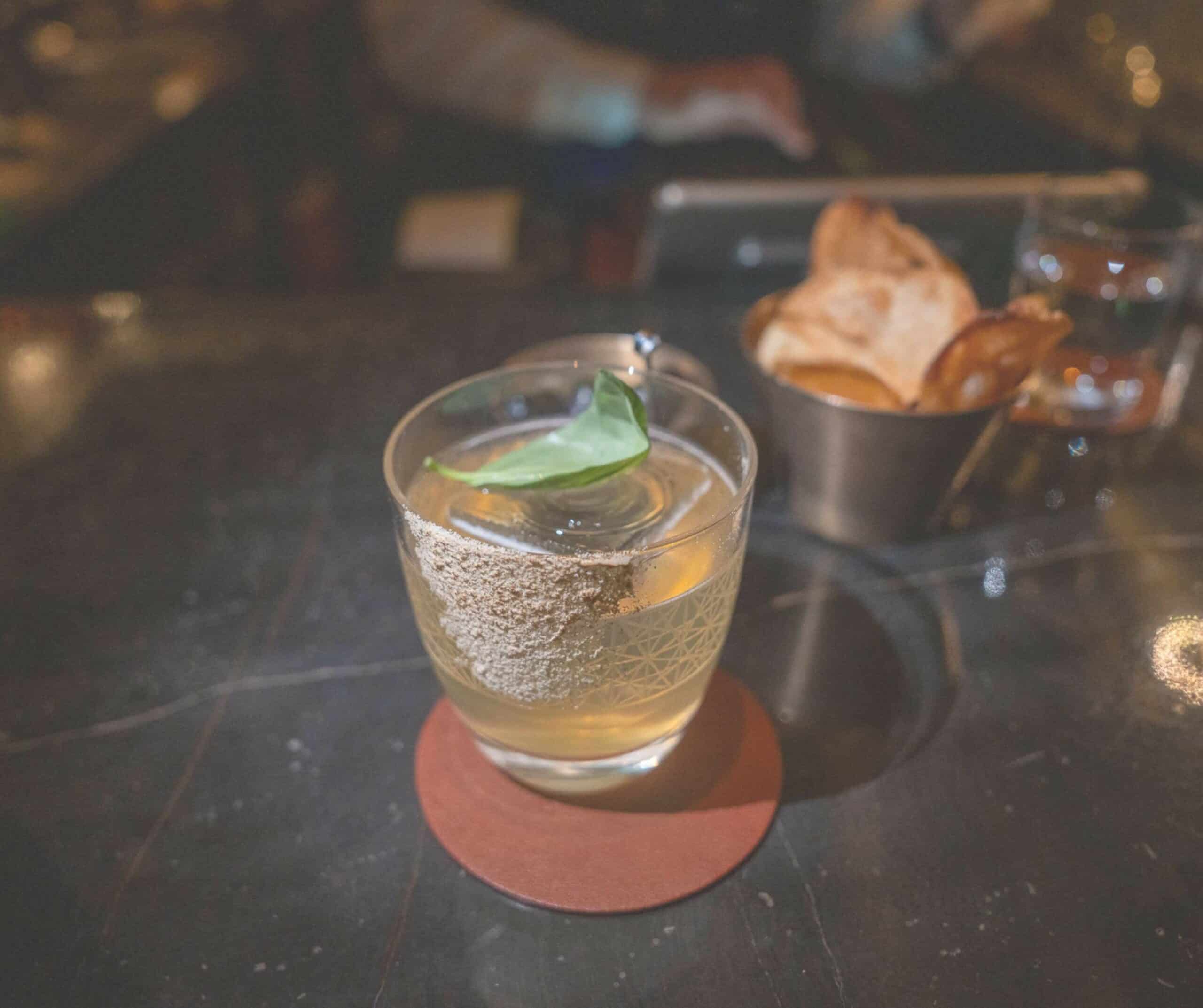 Cocktail in a sugar-rimmed glass with a mint leaf garnish on a red coaster at a dim bar.
