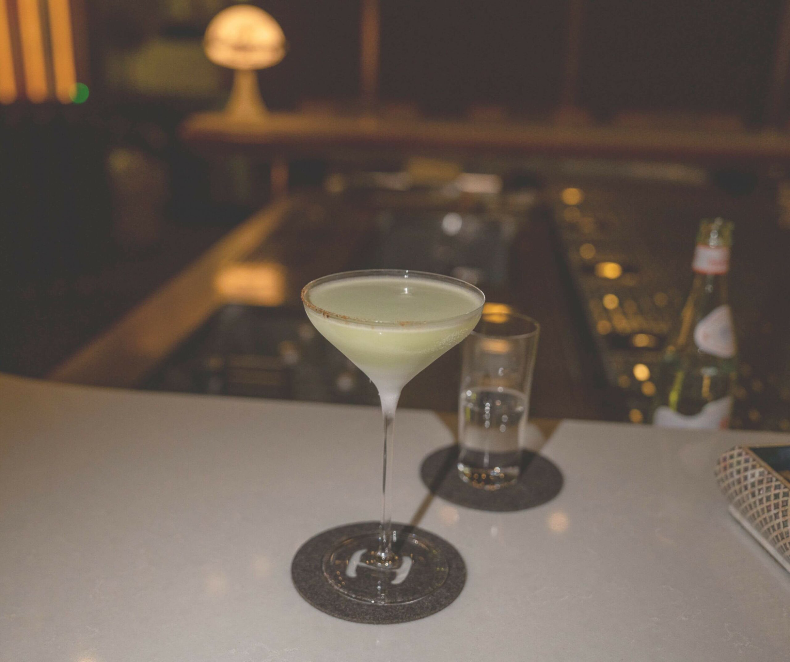 Blurred bar scene with a pale green martini in the foreground on a counter and soft lights in the background.