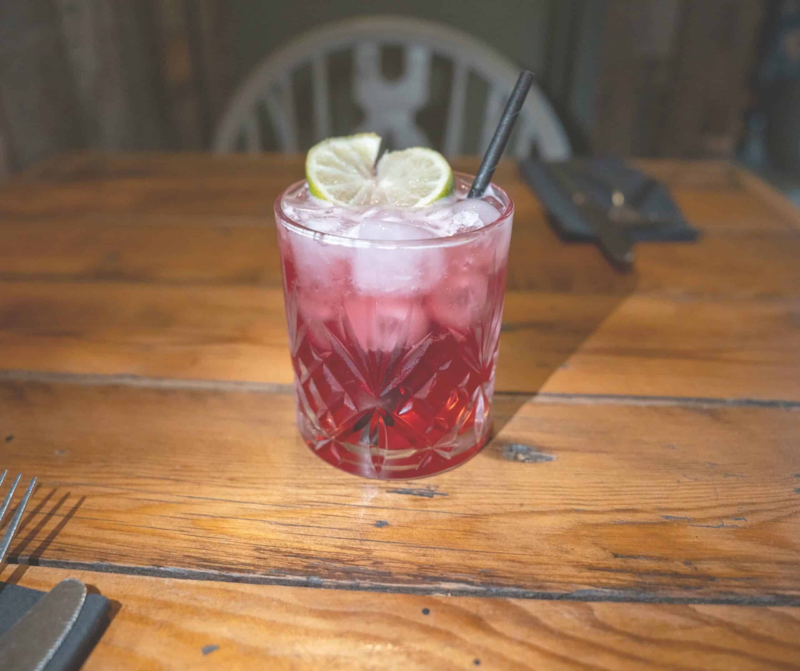 Pink cocktail with crushed ice and two lime wheels in a cut-glass tumbler on a wooden table at Hoof in Rye