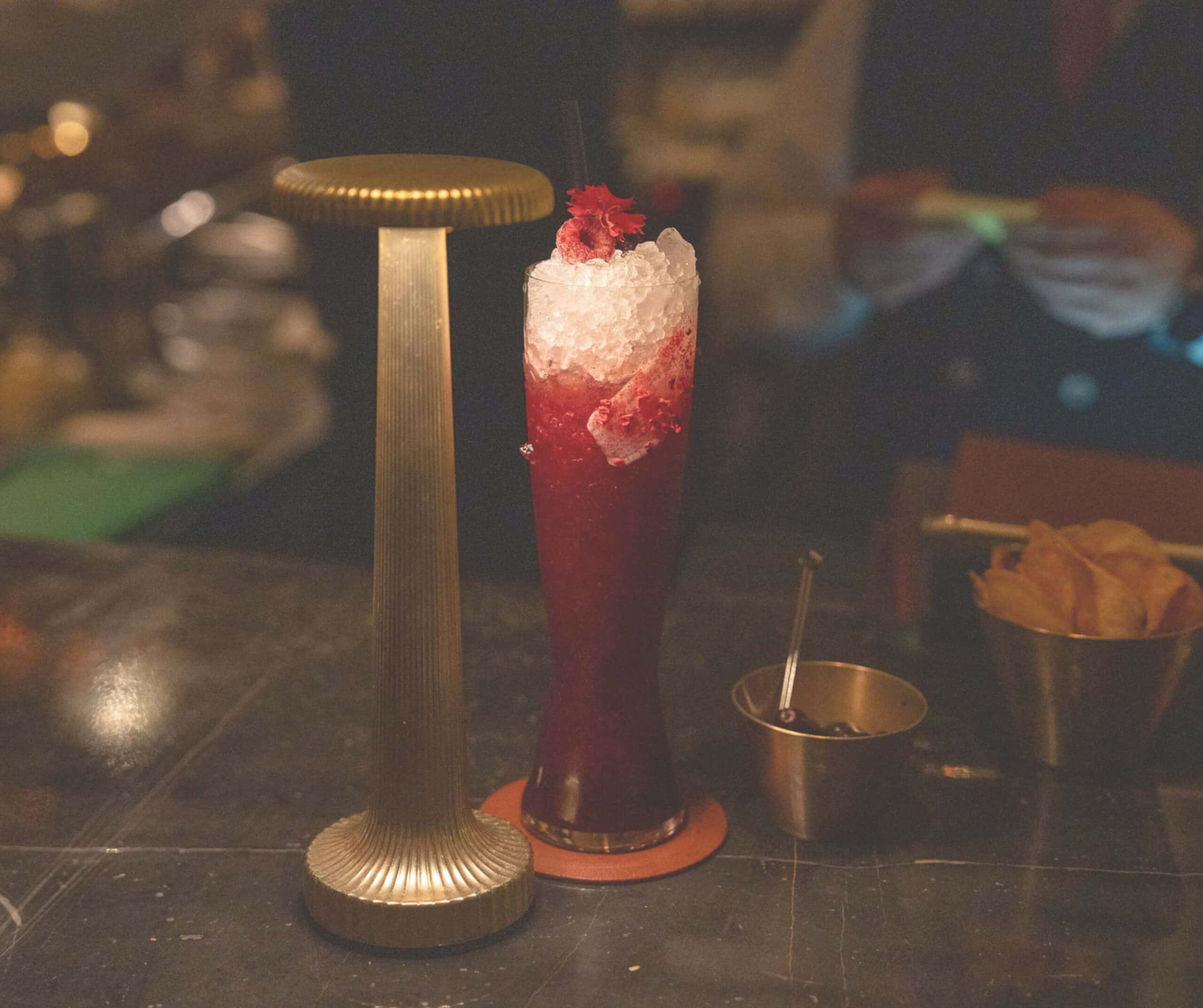 Tall red cocktail with crushed ice and a raspberry garnish on a coaster beside a brass bar lamp in a dimly lit setting, with a straw in the drink.