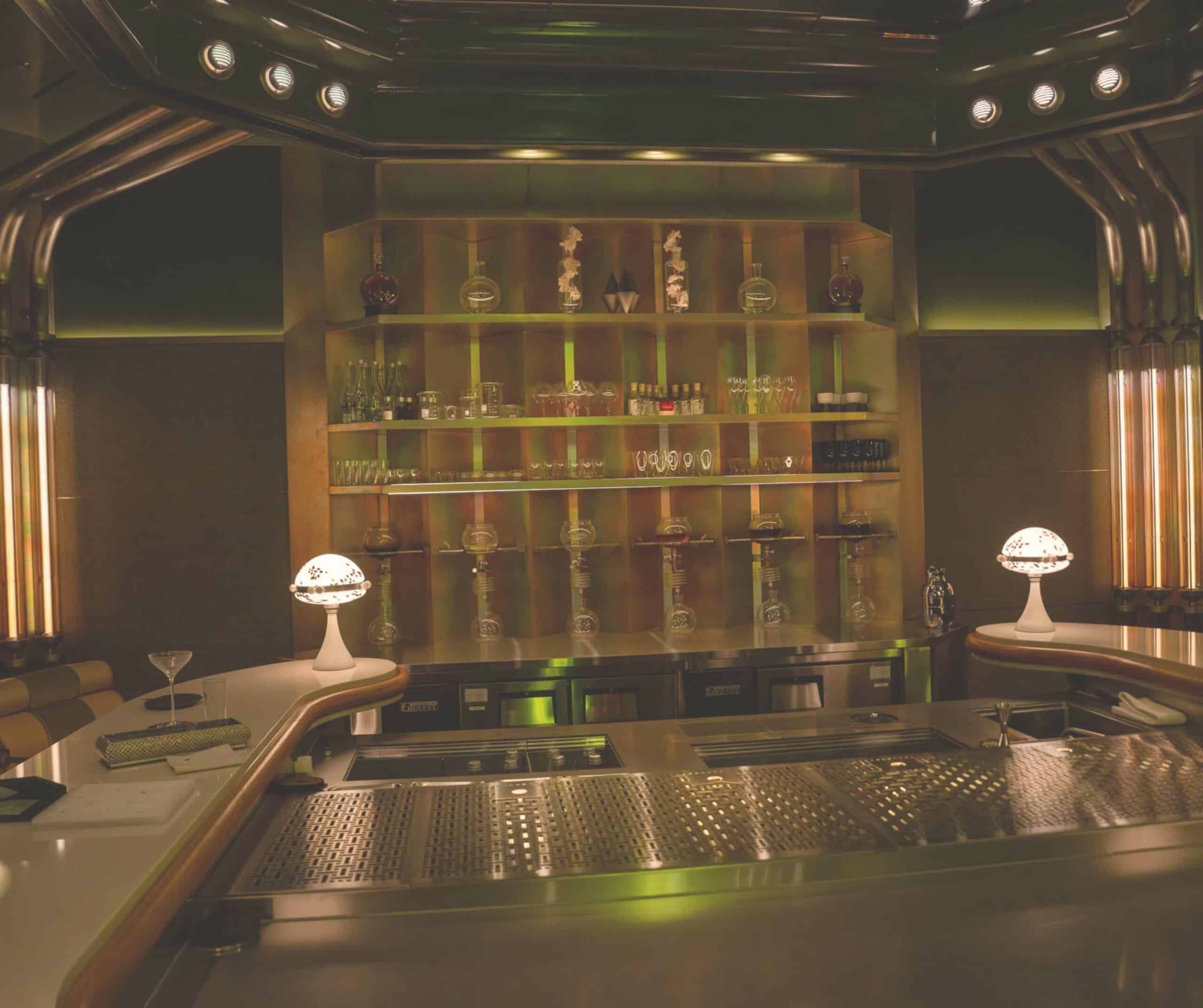 Dimly lit bar interior with metal shelves displaying liquor bottles, glassware, and decorative glass vases under warm lighting, in a retro style.