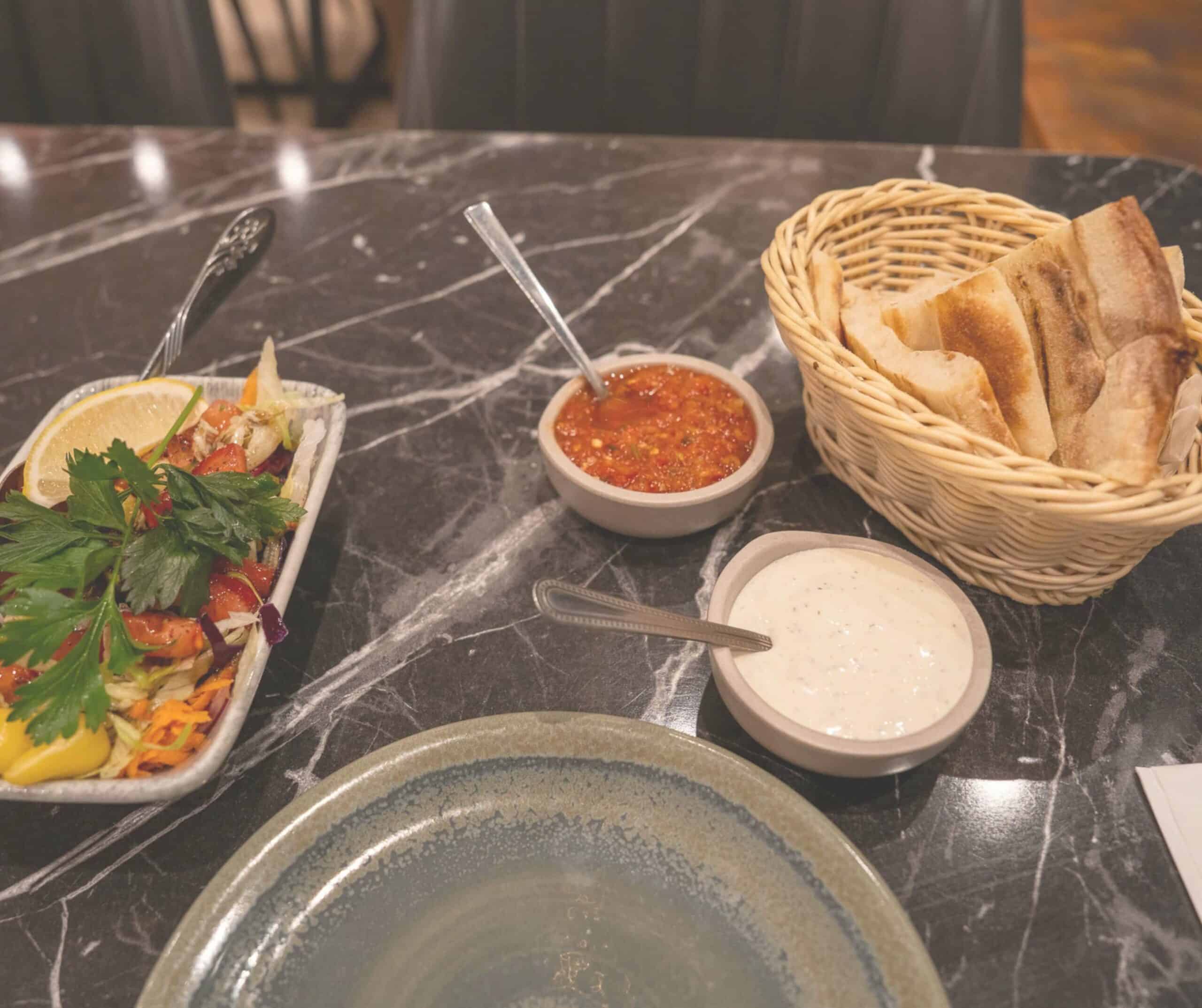 Complimentary warm pita bread, salad, and red and white dips on a marble table at Diamond Köz in Rye