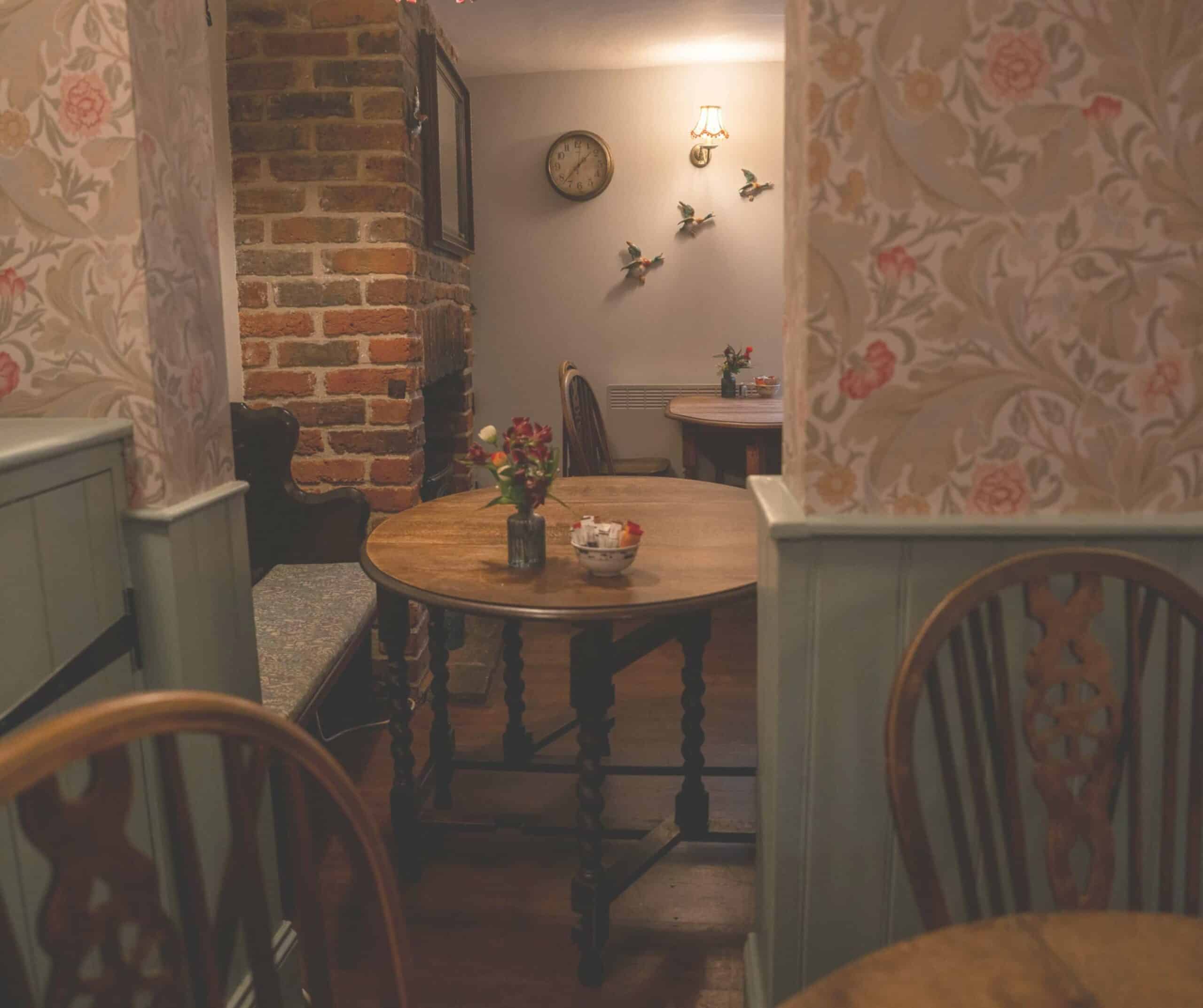 Cozy corner of The Cobbles Tea Room in Rye with floral wallpaper, a round table, vintage clock, and flying duck wall ornaments