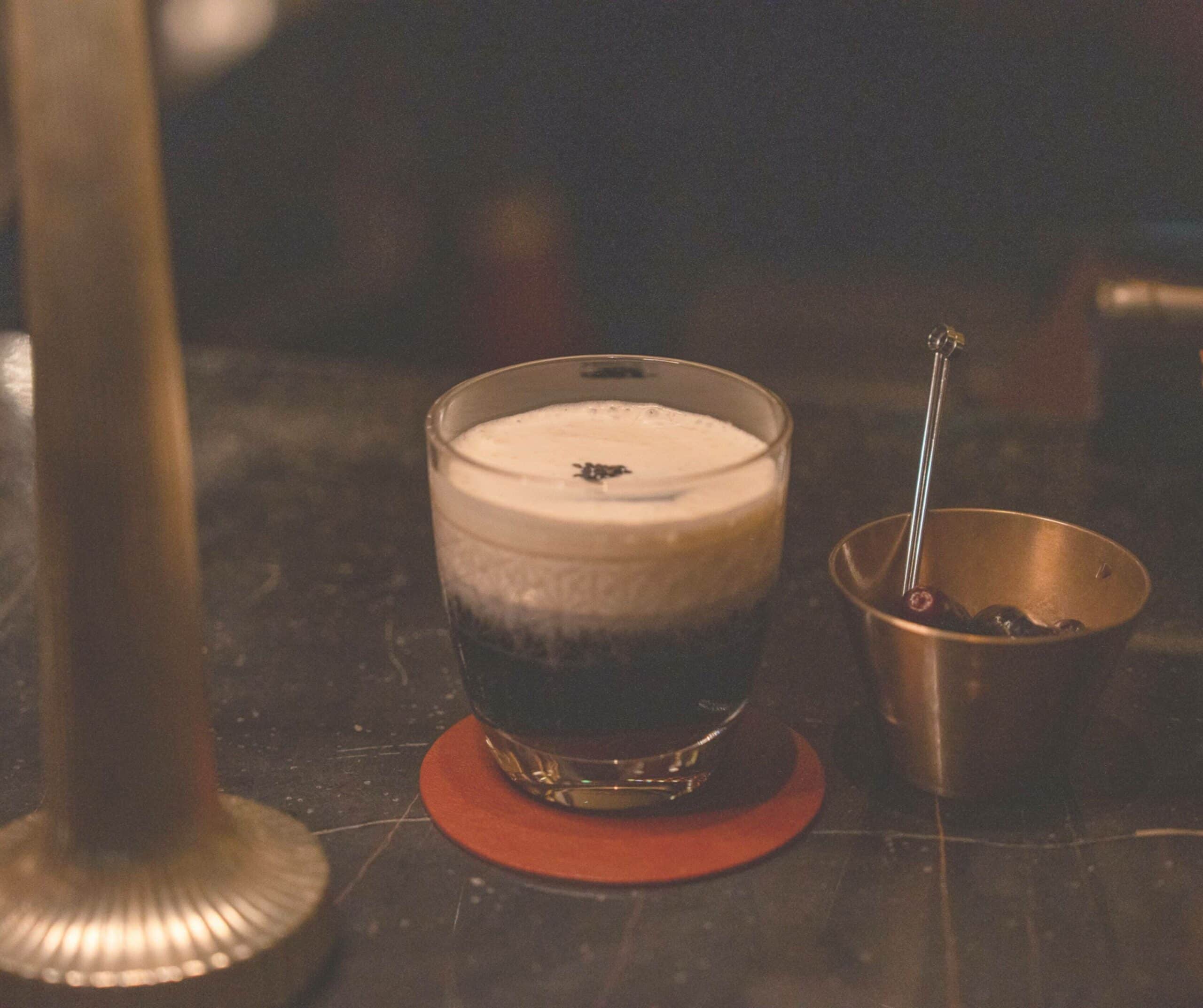 Two drinks sit on a dark bar counter; one has a foamy top and a metal stirrer nearby on an orange coaster.
