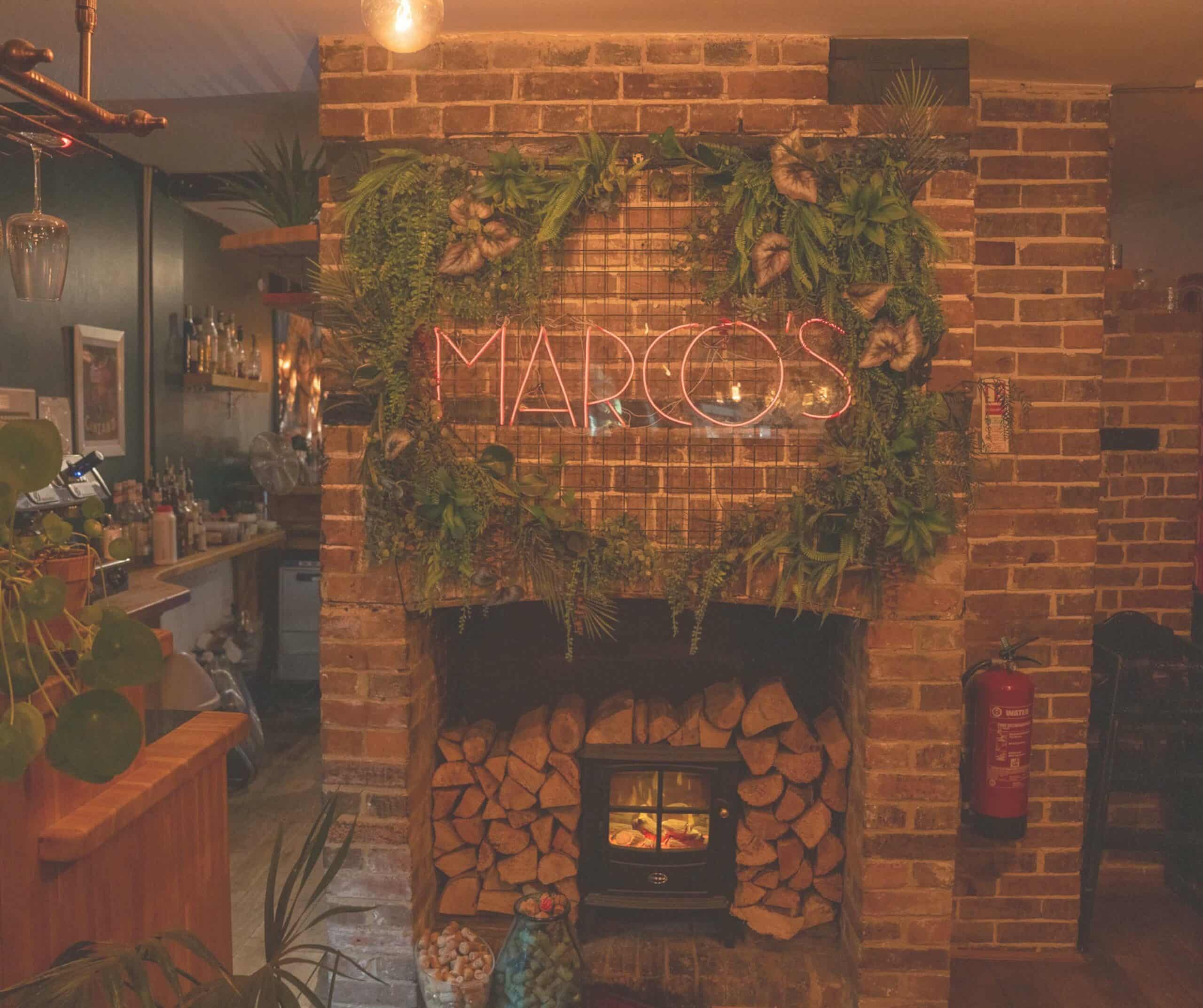 Brick fireplace with a log burner, hanging plants, and a pink neon Marco's sign at Marco's Mediterranean Kitchen in Rye
