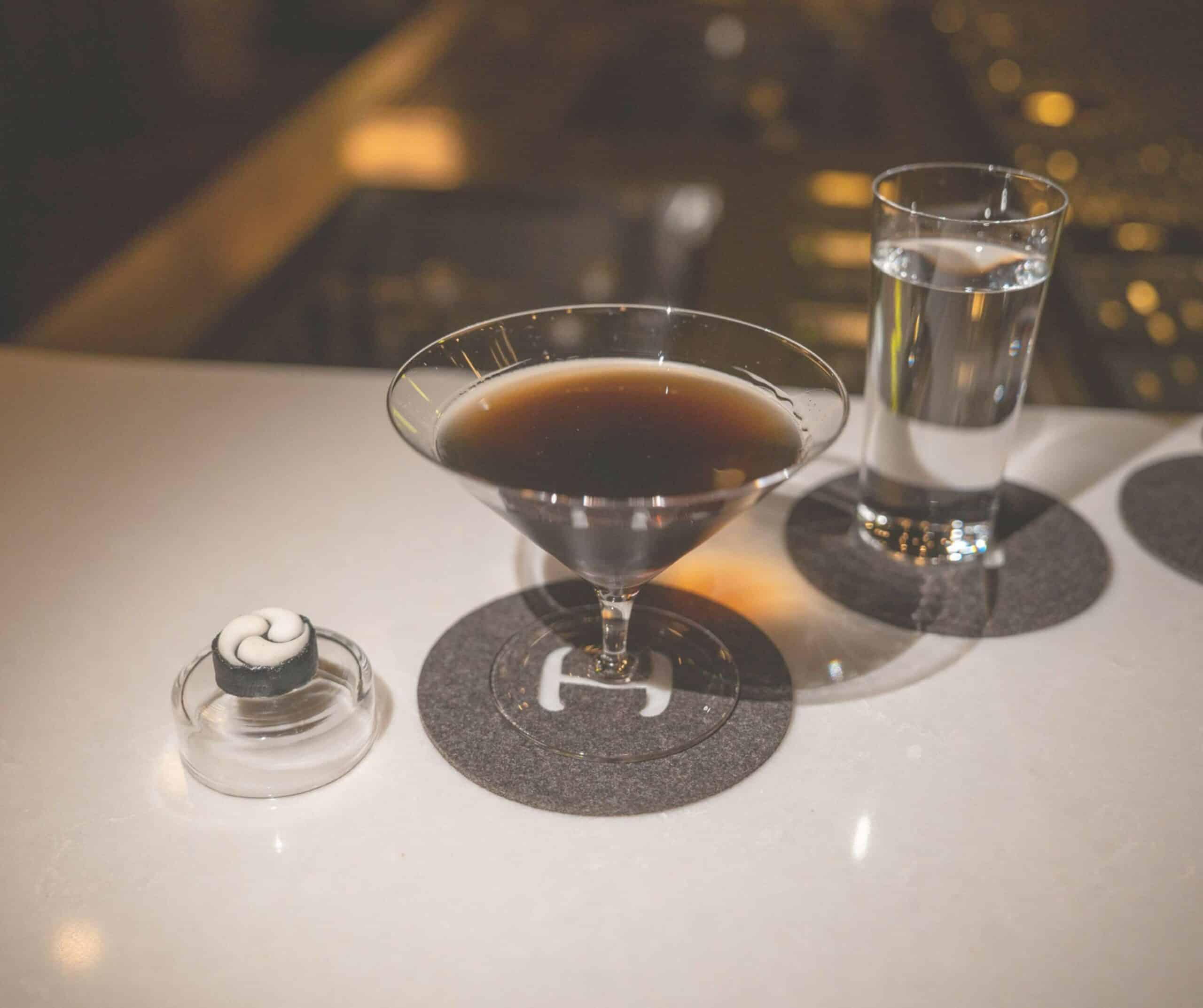 Close-up of a dark cocktail in a martini glass on a white table, with a tall glass of water and a small dessert in a separate glass, against a blurred nighttime background.