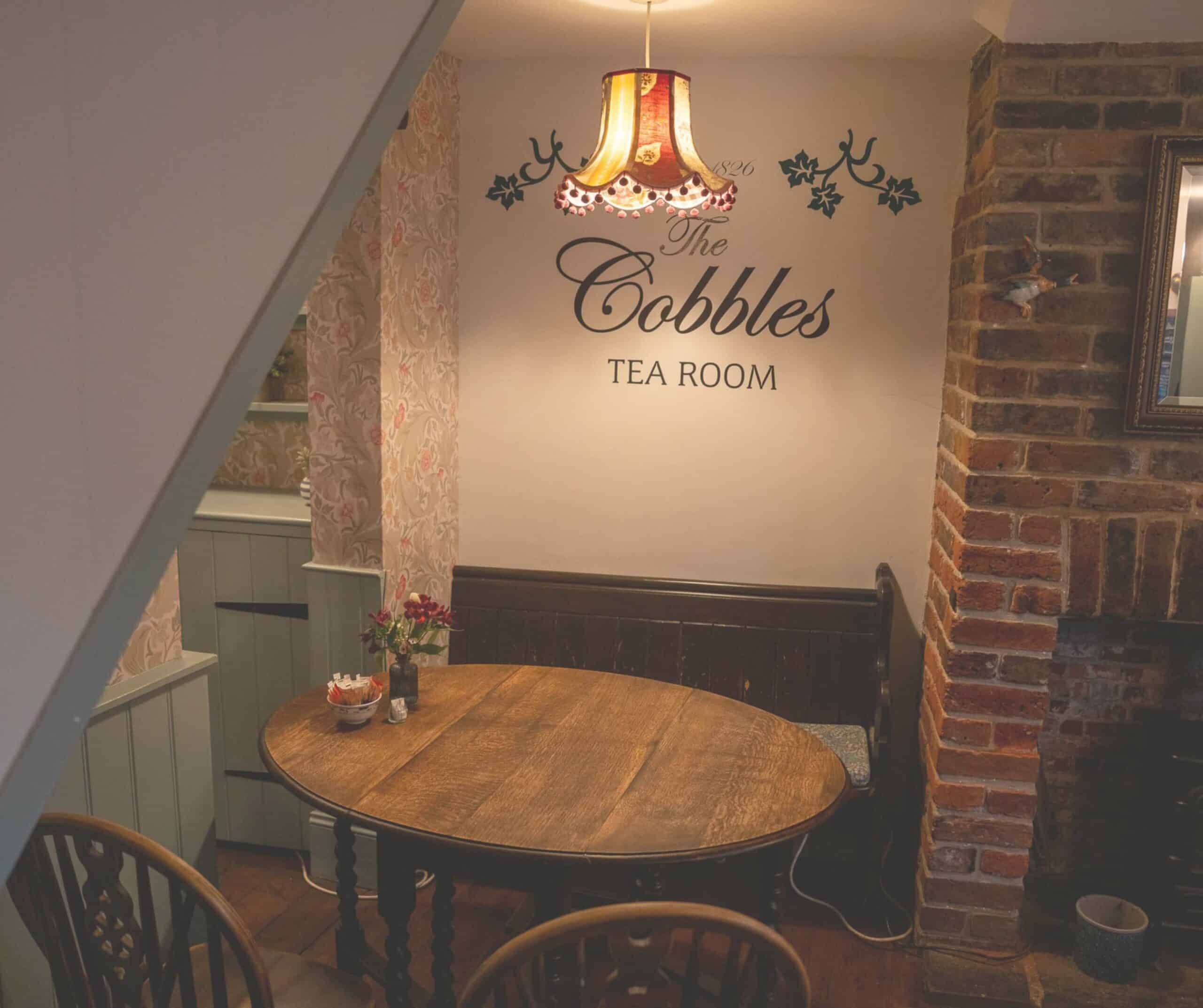 The Cobbles Tea Room sign painted on the wall in Rye above a wooden table, bench, and exposed brick chimney