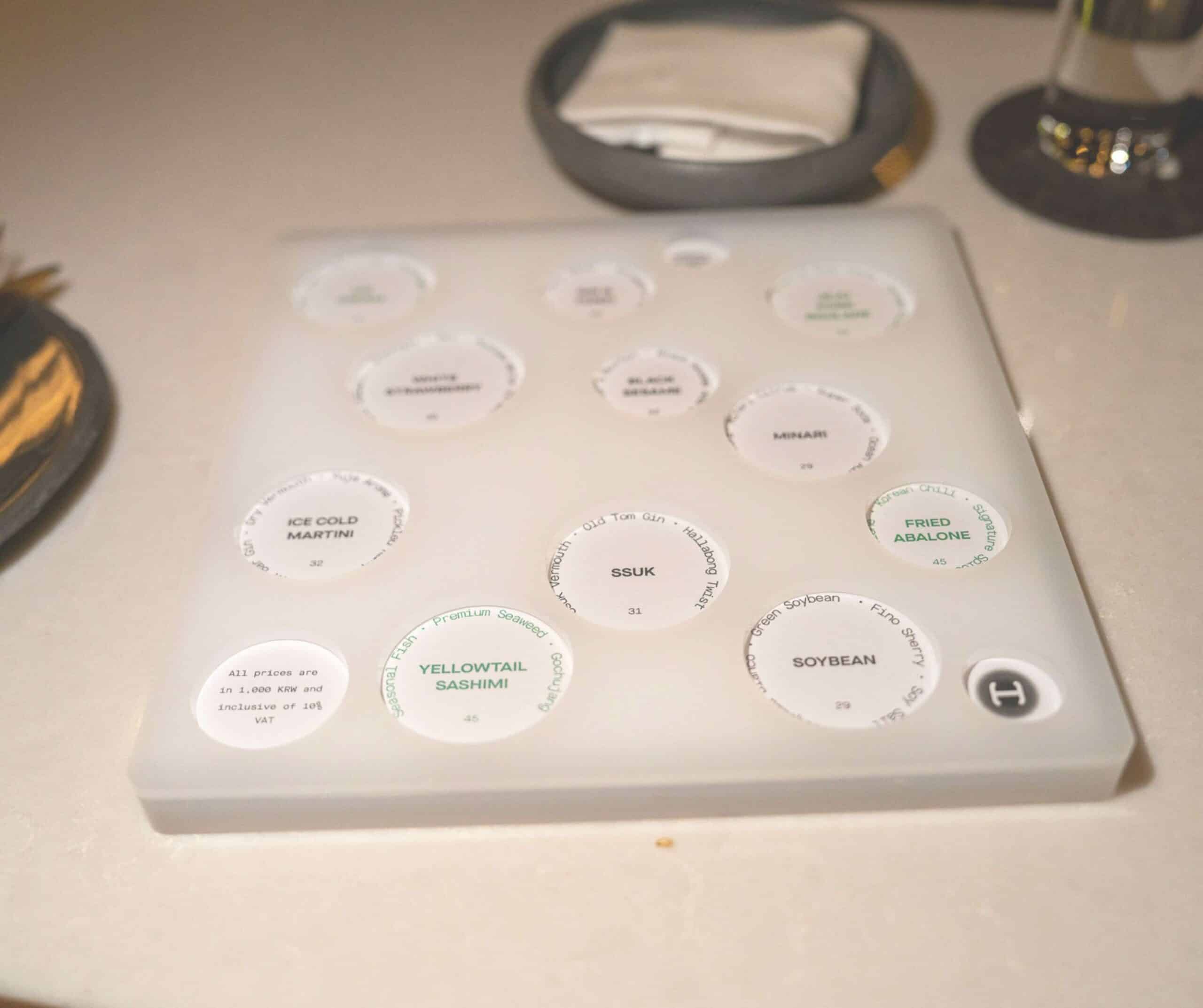 Close-up of a white tray with round dish labels listing sushi items like Yellowtail Sashimi and Fried Abalone.